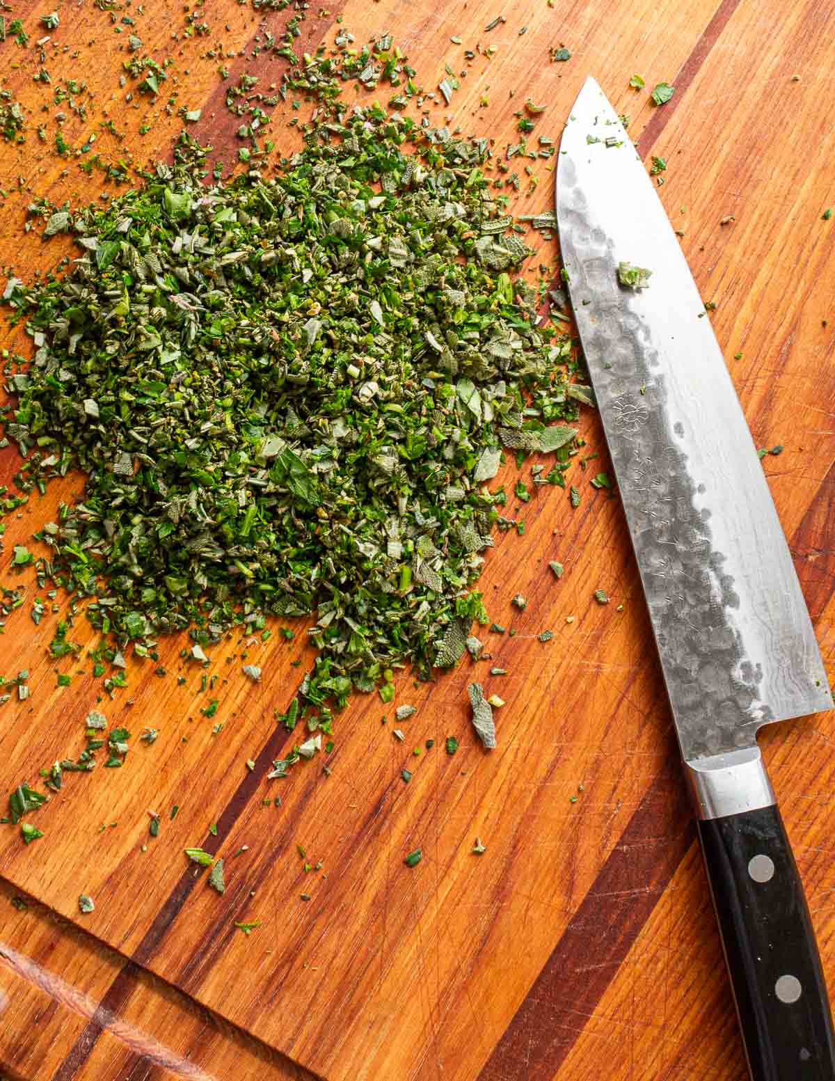 Finely chopped rosemary sage and thyme on a cutting board next to a knife.