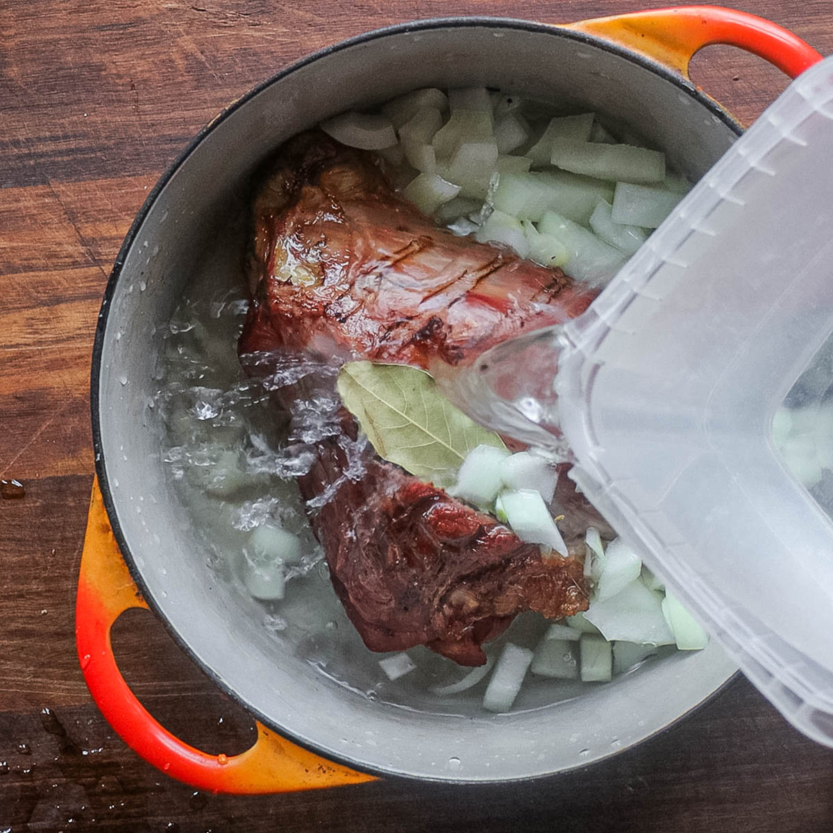 Adding water to a pot of smoked lamb shank and onions.