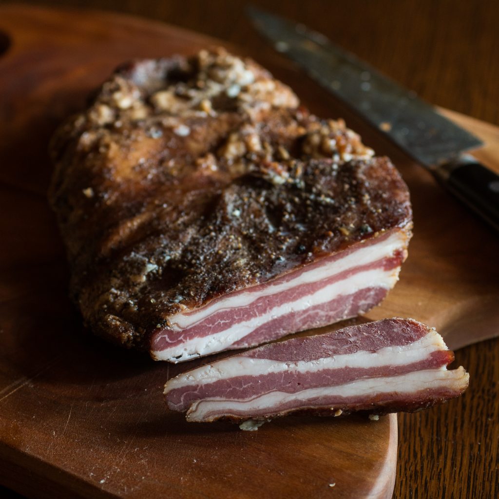 A slab of cooked lamb bacon sliced revealing the pink inside on a cutting board next to a knife. 