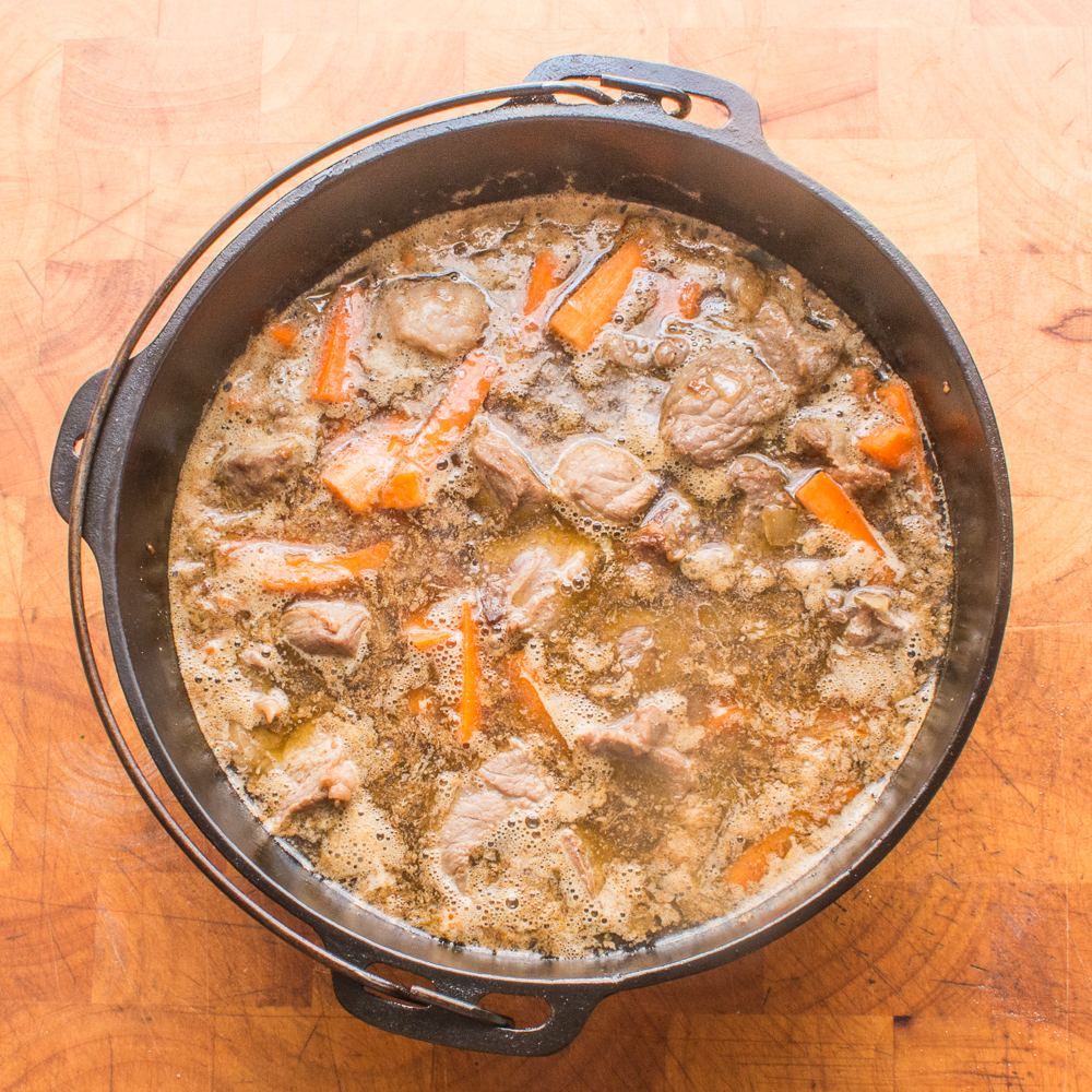 Adding browned lamb meat and stock to carrots and onions in a Dutch oven.