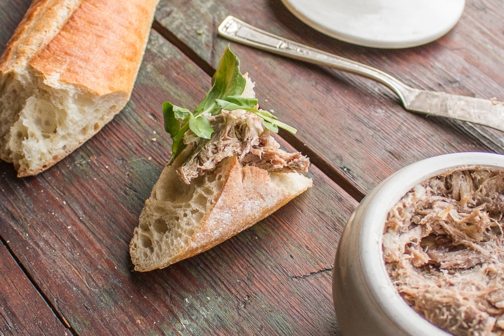 A bageutte with rillettes on top next to a pot full of rillettes. 