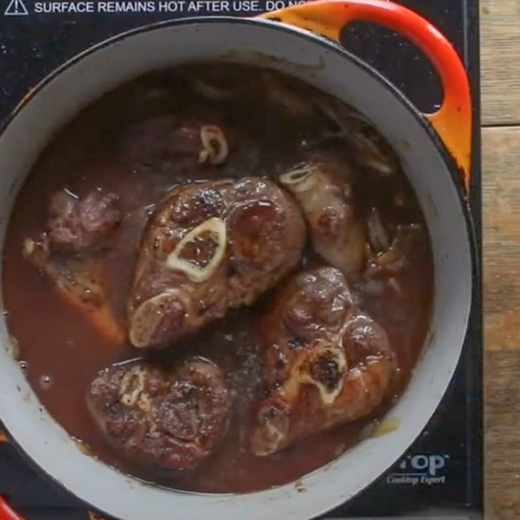 Adding stock to a pot of cooking lamb shanks.