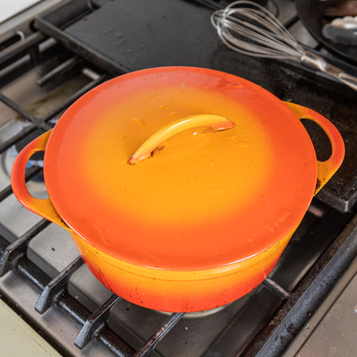 An orange enameled cast iron dutch oven with the lid on cooking over a burner.
