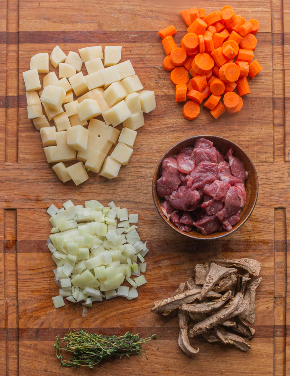 A top down image of lamb stew meat ingredients chopped up. Potatoes, carrots, onion, lamb meat, dried mushrooms and thyme.