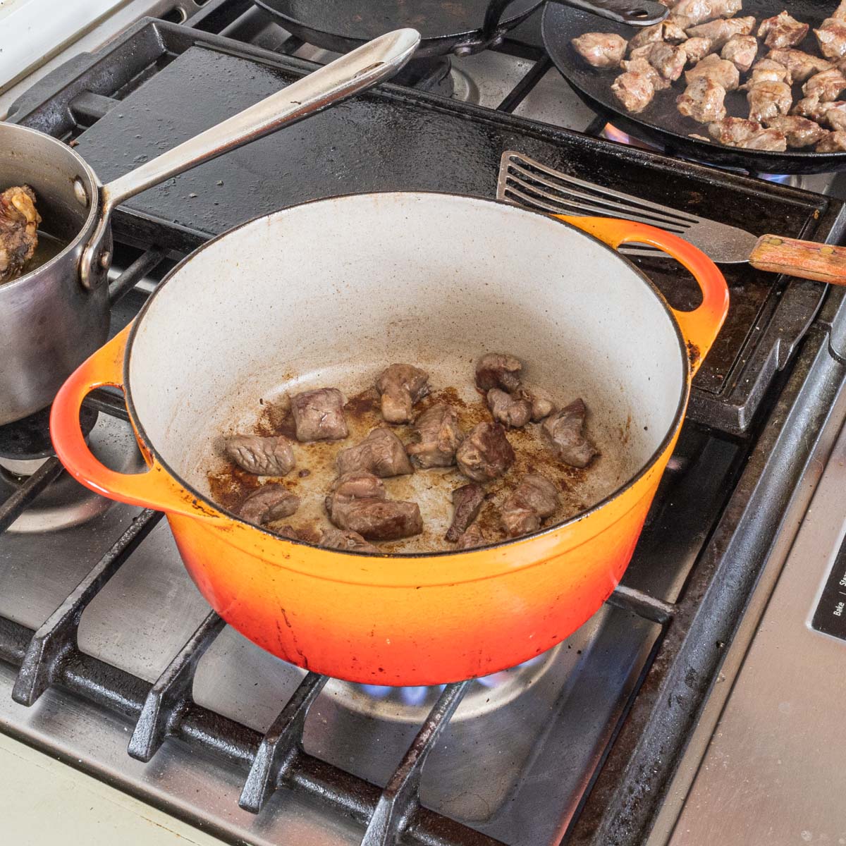 Browning lamb meat stew pieces in an orange enameled cast iron dutch oven on a burner.