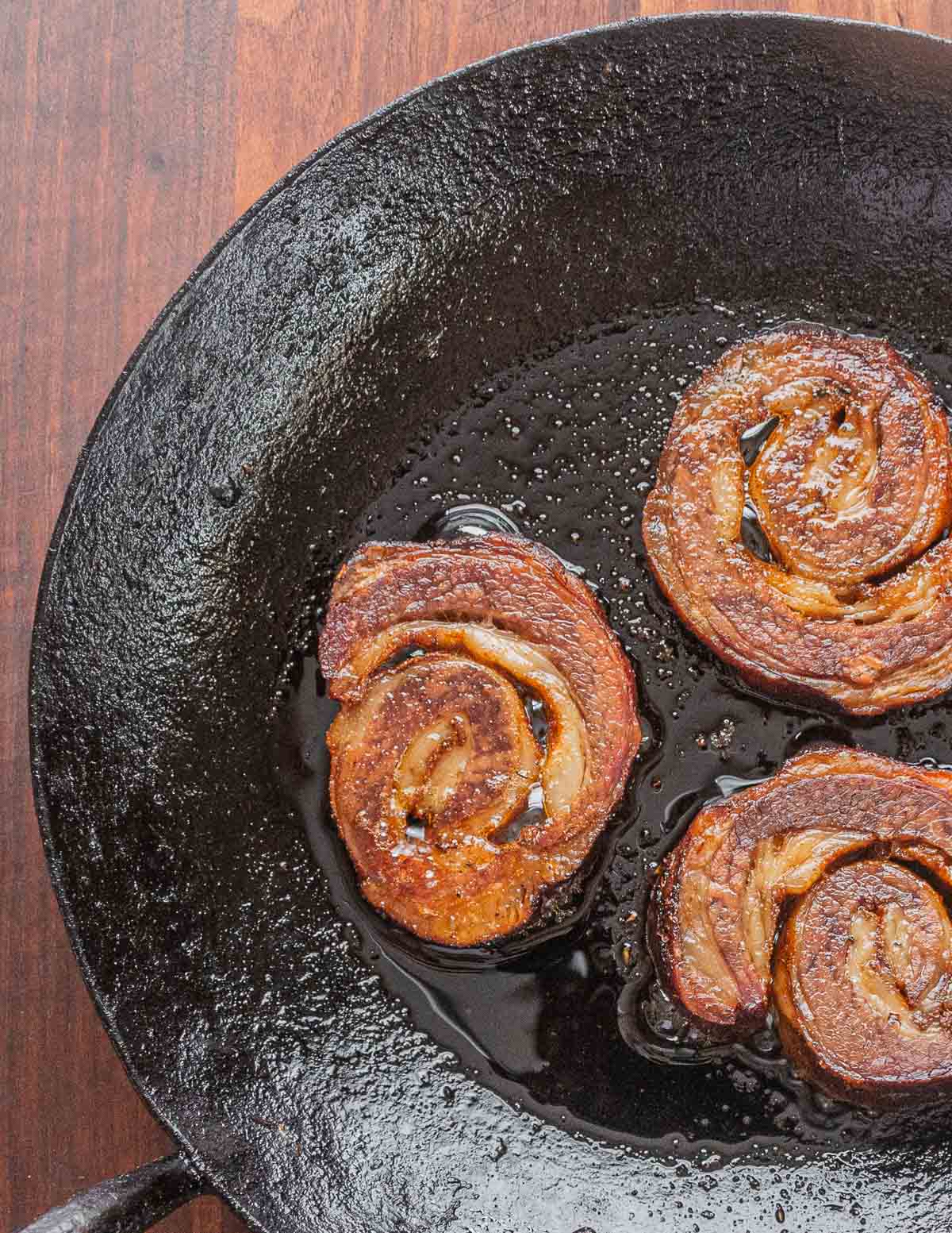 Rolled slices of smoked lamb breast cooking in a pan.
