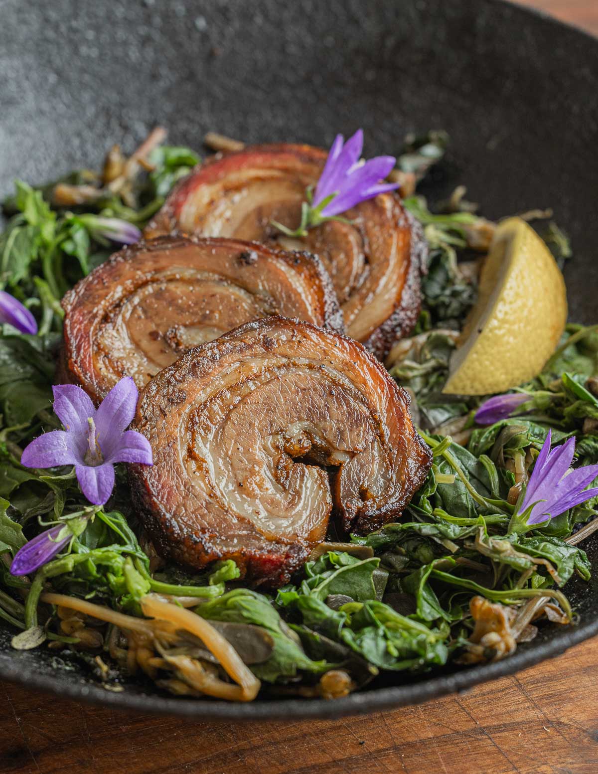 Sliced browned smoked lamb breast in a pan with cooked greens, bellflowers and lemon. 