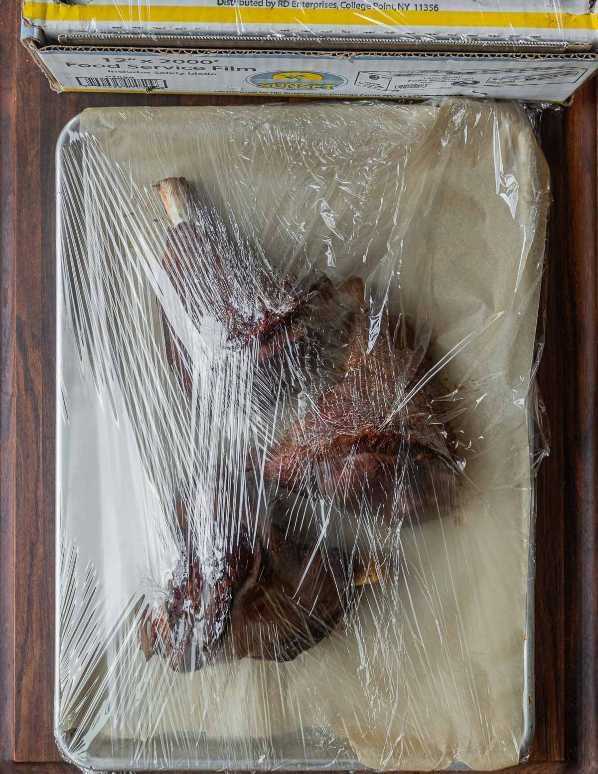 Smoked lamb shanks on a tray lined with parchment wrapped in cling film.