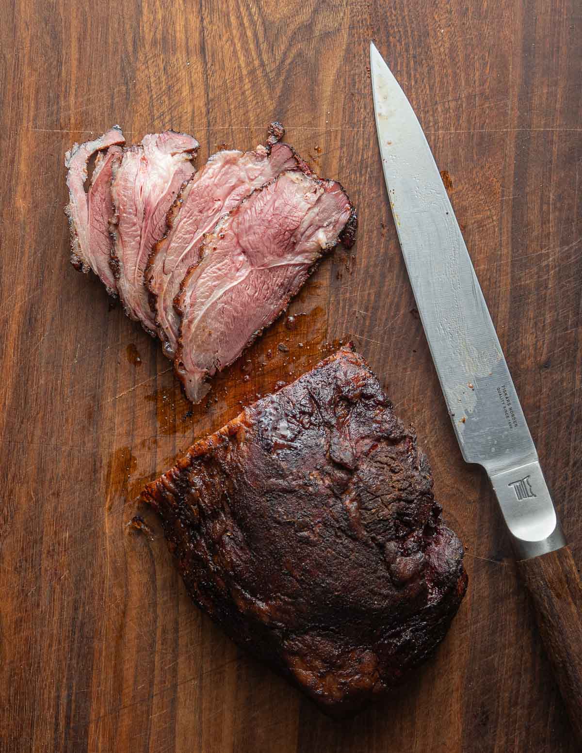 A pink or medium temperature smoked lamb leg sliced with a long knife on a black walnut cutting board.