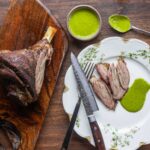 A smoked lamb shank carved on a cutting board and served sliced ona plate with green sauce.
